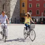 Persone in bicicletta in piazza soleggiata davanti caffè.